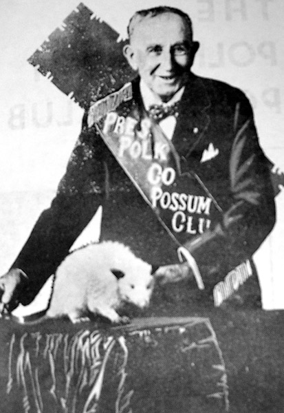 white man in sash "Pres. Polk Co. Possum Club" poses with an opossum