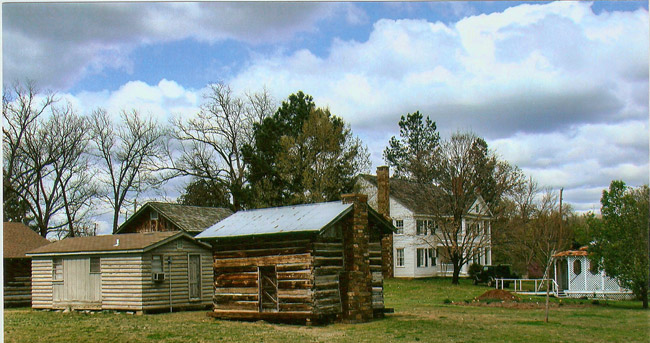 Single-story cabin and outbuildings with multistory house with covered porch and balcony in the background