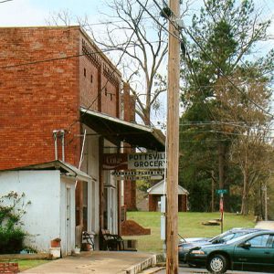 Side view of brick storefront with covered sidewalk and signs on street with parked cars and multistory house in the background
