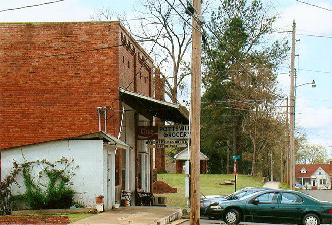Side view of brick storefront with covered sidewalk and signs on street with parked cars and multistory house in the background