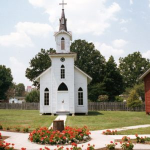 Courtyard including white wood frame church and separate red school building with bell tower and walkways with red flowers