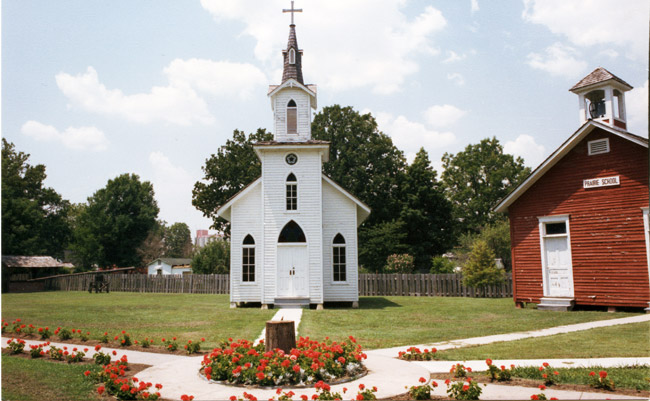 Courtyard including white wood frame church and separate red school building with bell tower and walkways with red flowers