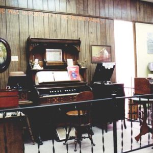Interior museum displays behind railing including piano phonograph antique furniture and description "Music Was Important in the Prairie Home"