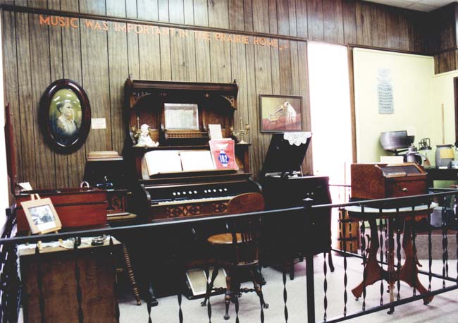 Interior museum displays behind railing including piano phonograph antique furniture and description "Music Was Important in the Prairie Home"