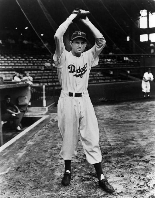 White male baseball player "Dodgers" uniform poses arms glove raised few players spectators watching