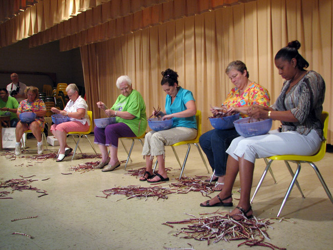 Row of women shelling purple hull peas seated on plastic chairs stage curtains in background