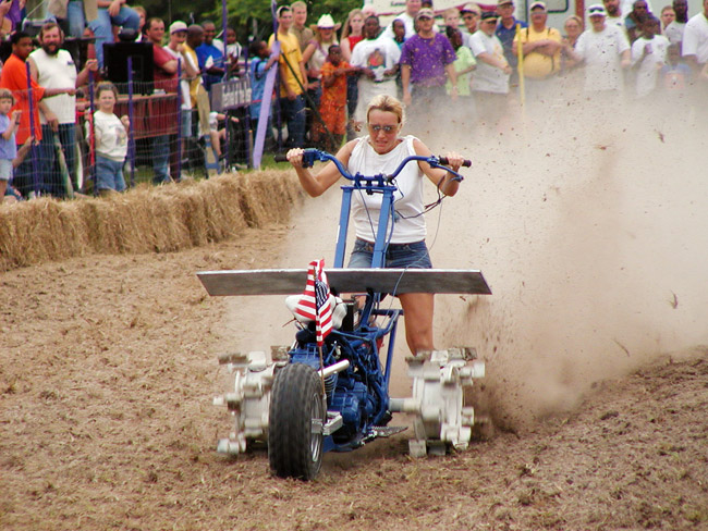 White woman riding motorcycle tiller on dirt track with hay bales fence audience