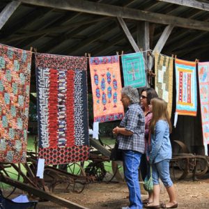 women looking at colorful quilts hanging on clothes line in a barn