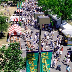 Aerial view of crowd on paved parking lot with striped tents and multistory buildings in the background