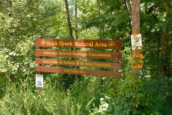 "Rock Creek Natural Area Arkansas Natural Heritage Commission" sign surrounded by trees and green foliage