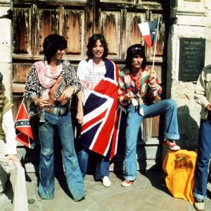 five white men, one draped in British flag and one waving Texas flag, outside weathered wooden doors