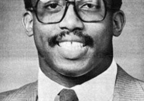 Portrait of a black man in a suit, tie, and glasses.