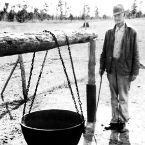 White man in hat and jacket with large pot suspended by chains from a log