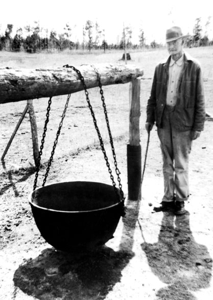 White man in hat and jacket with large pot suspended by chains from a log