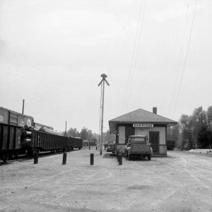 Gravel lot with tall pole between freight trains and a small "Sheridan" station with truck parked by it