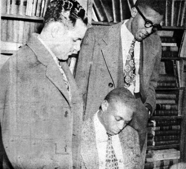 Two black men standing and one seated in library wearing suits jackets and ties gazing downwards