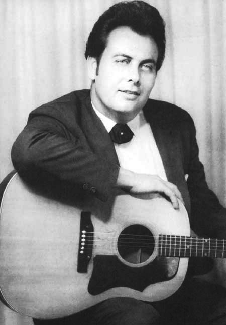 White man in suit and bow tie with dark pompadour and sideburns seated smiling with his arm on acoustic guitar