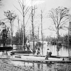 Three men with pole boats transporting iron pipes in flooded area with tall trees near oil derrick.