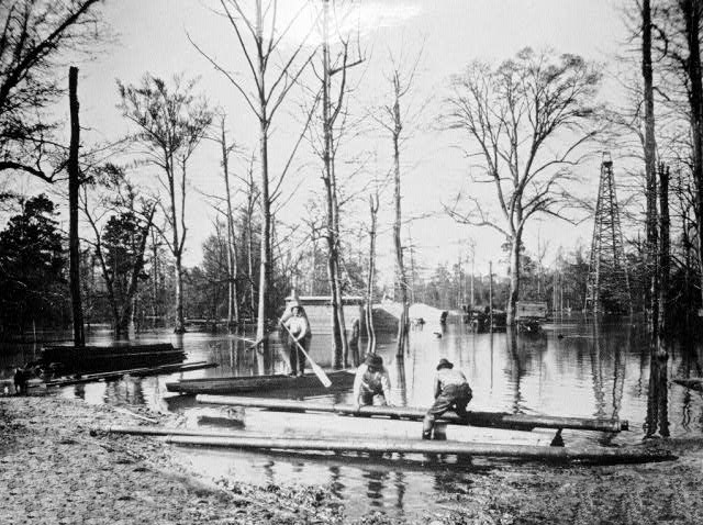 Three men with pole boats transporting iron pipes in flooded area with tall trees near oil derrick.