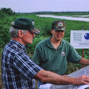 Older white man in hat and plaid shirt with younger white man in hat and green shirt in wetlands with map on car hood