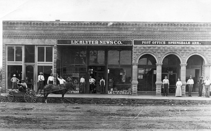 Exterior stone and brick building with sign "Lichlyter News Co.," "Post Office Springdale Ark." with onlookers and horse-drawn carriage