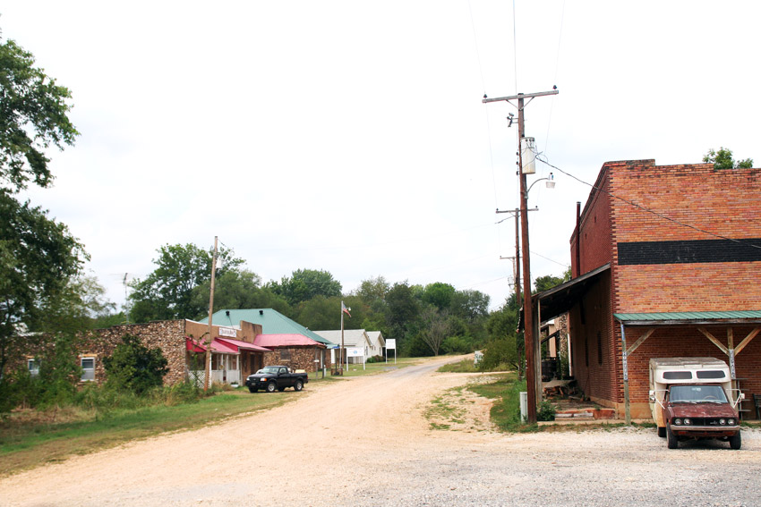 Dirt road with brick and stone storefronts on the left side and multistory buildings with covered porches on the right side