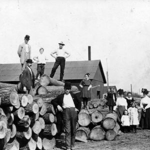 White men, women, and children pose on cut tree lumber near train tracks barn-like structures in background