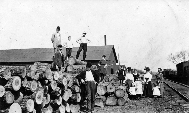 White men, women, and children pose on cut tree lumber near train tracks barn-like structures in background