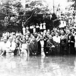 Group portrait white men and women standing in river with onlookers on tree-lined riverbank