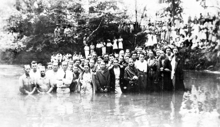 Group portrait white men and women standing in river with onlookers on tree-lined riverbank