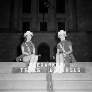 Two young white women in matching hats and dresses sitting on stairs with Texas-Arkansas border line painted on them and stone building in background