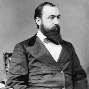white man with beard in suit and tie seated by small stack of books