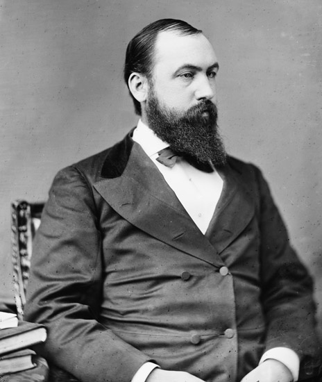 white man with beard in suit and tie seated by small stack of books