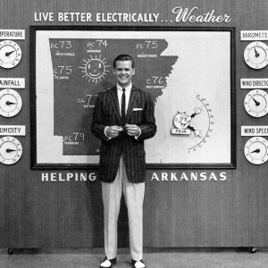 White man in stripped suit jacket poses with Arkansas weather map