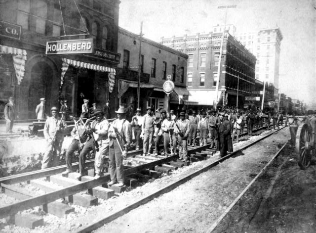 Black railroad workers stand on tracks downtown in perpendicular groups of four holding picks and hammers