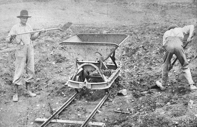 men in hats boots and pants with suspenders loading a mine cart with shovels