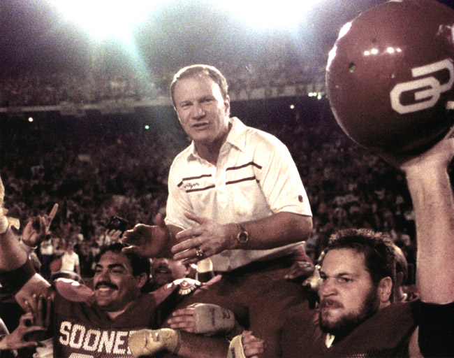 White football coach hoisted on "Sooners" players shoulders  with stadium crowd in background