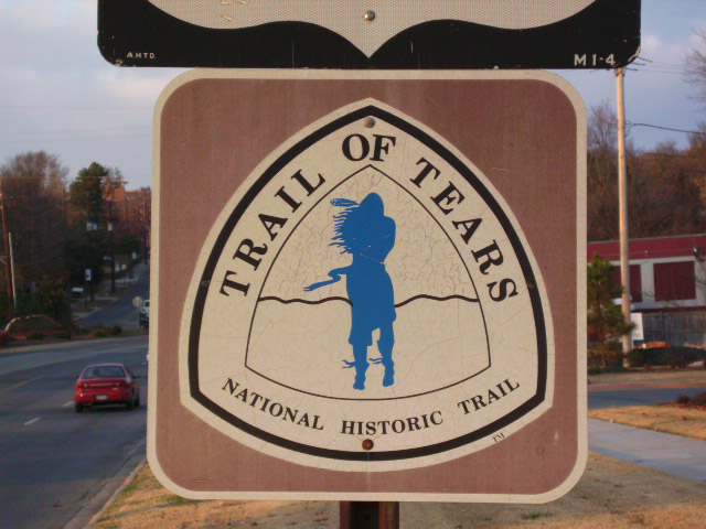 Street sign with triangular logo labeled "Trail of Tears National Historic Trail" featuring Native American man in its center in blue