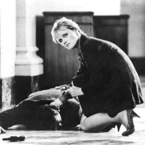 White woman kneeling and pointing a gun checking pulse of man lying face down on floor