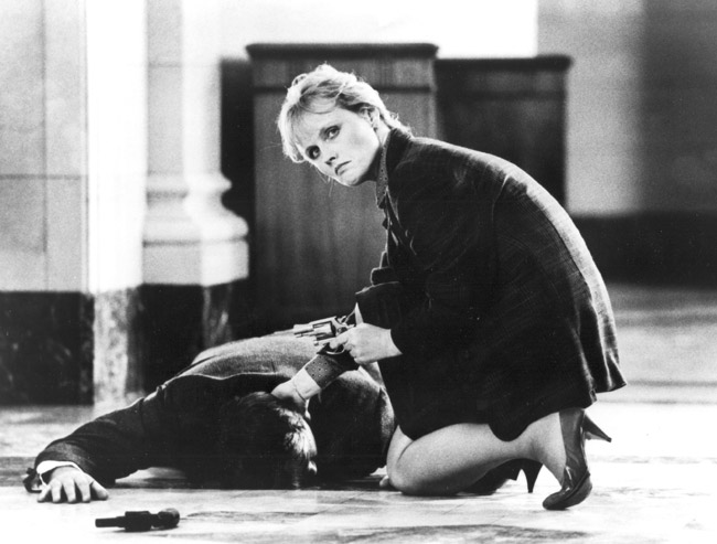 White woman kneeling and pointing a gun checking pulse of man lying face down on floor