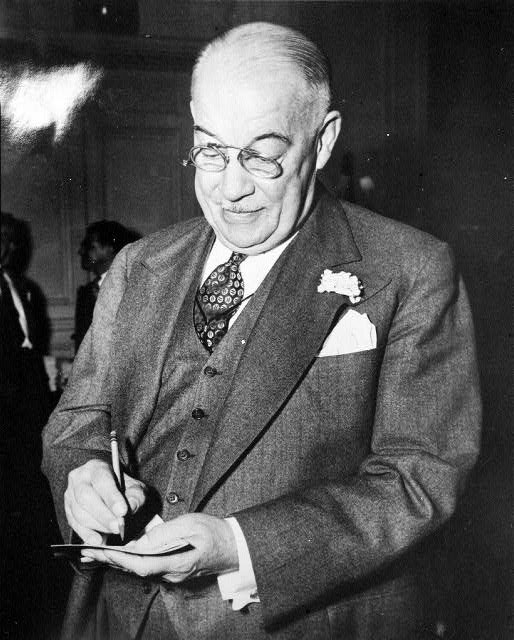 Older white man wearing glasses in suit and tie signing a piece of paper with a pencil