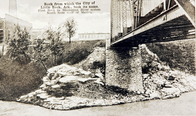 Underside of steel truss bridge over river with brick supporting columns and multistory buildings in the background with writing saying "rock from which the city of little rock Arkansas took its name"
