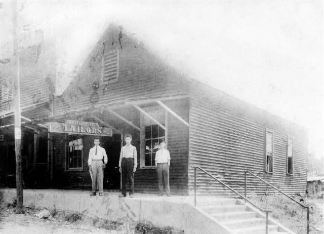 Three men in dress shirts stand on sidewalk below awning with "Tailors" sign near stairs