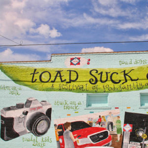 light blue wall with painted green banner reading "Toad Suck Daze a Festival of food, family, and fun" with paintings of events involved with festival