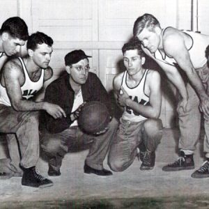 Young white men crouching in basketball uniforms with coach in black ball cap holding a basketball