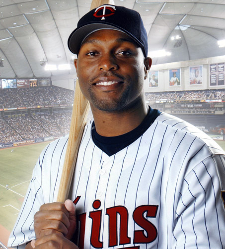 Young African-American man in striped uniform and cap posing with baseball bat over his left shoulder and stadium interior in the background behind him