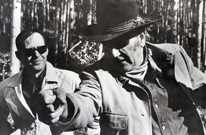 Two white men in aspen grove with man in cowboy hat and eyepatch making pistol grip gesture