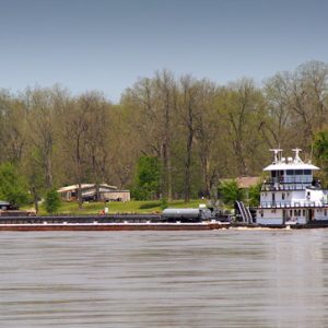 tug boat with barge on river with trees and structures on shore