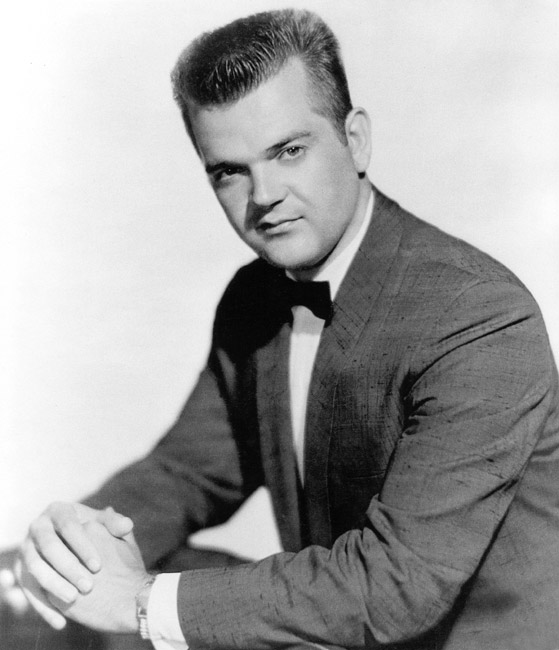 White man seated with hands crossed and a small smile with short hair combed upwards wearing suit jacket and bow tie