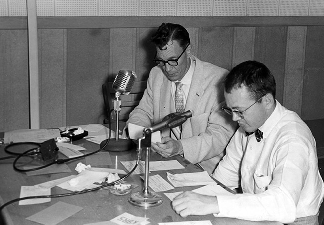Two white men with glasses reading at table with microphones and pieces of paper on the table